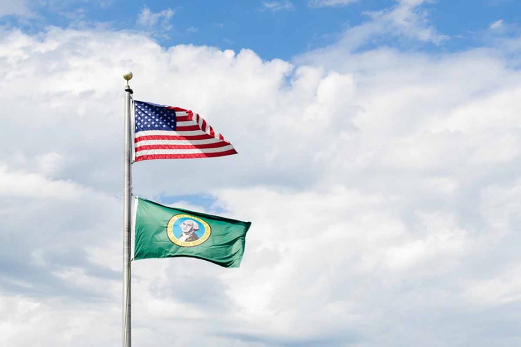 Flags of Washington state and the United States flying against clouds and blue sky.